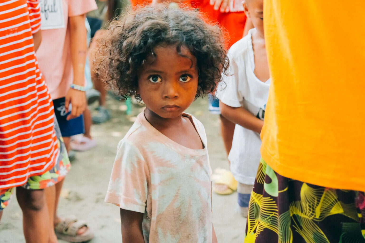 A young child looks straight into the camera, standing among volunteers and neighbours during the outreach