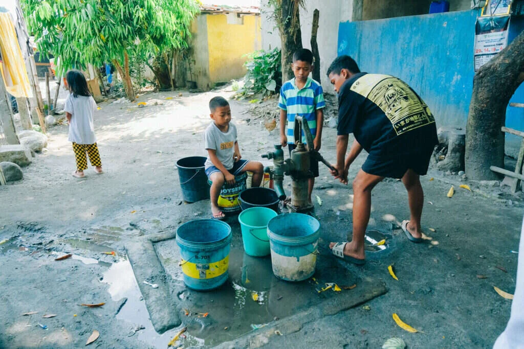 Children pumping water from a hand pump beside blue-painted homes